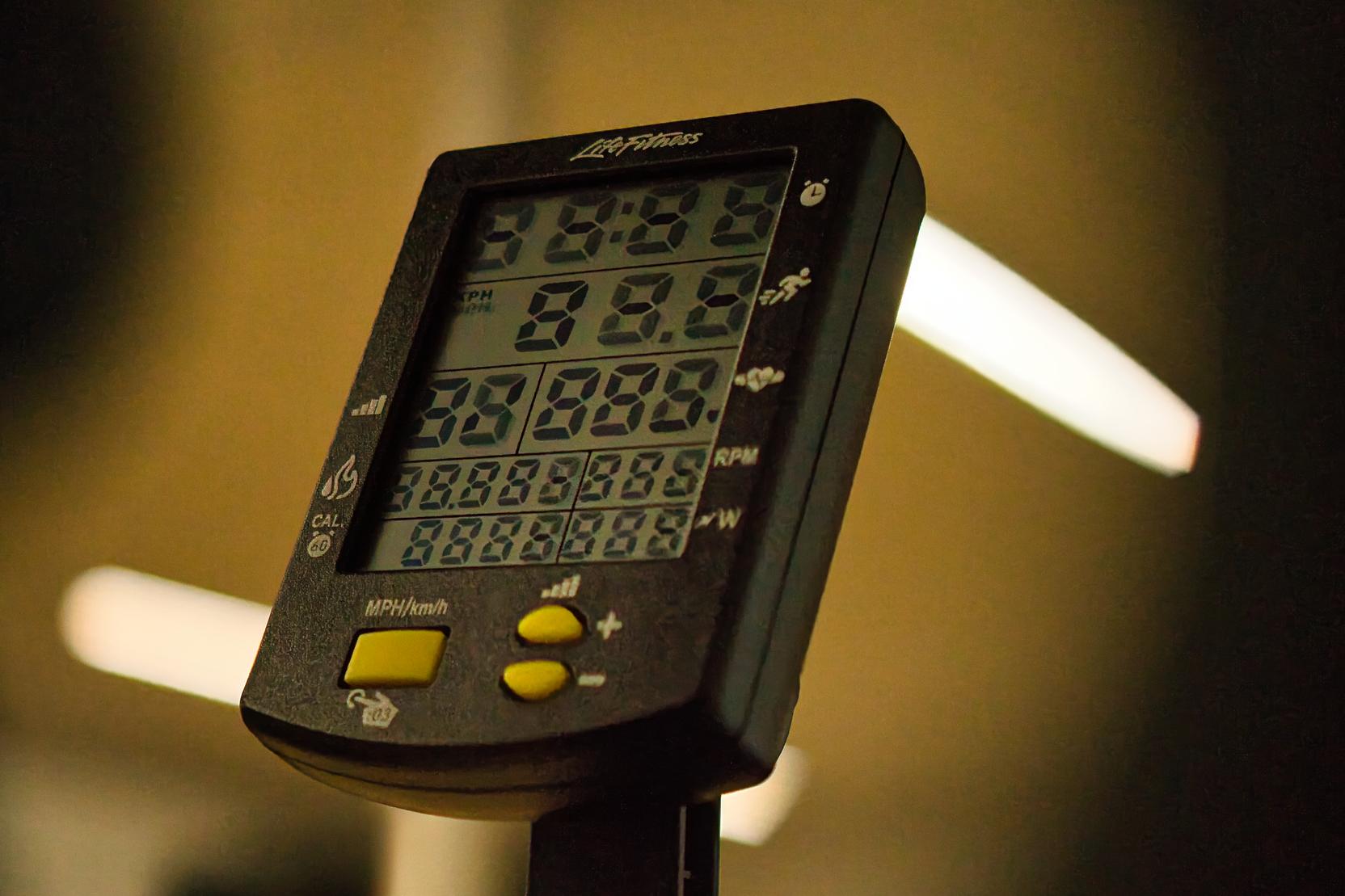 Display eines Fitnessger&auml;ts mit digitalen Werten wie Zeit, RPM und Watt, unscharf im Vordergrund.