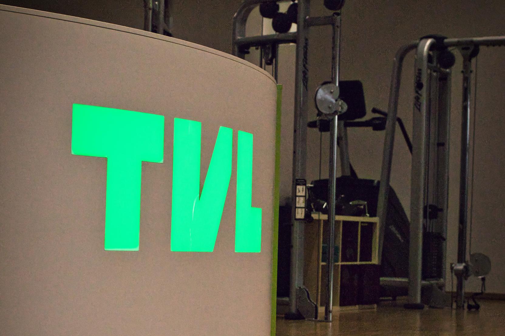 Beleuchteter Empfangstresen mit der Aufschrift "TVL" in gr&uuml;n, im Hintergrund verschiedene Fitnessger&auml;te.