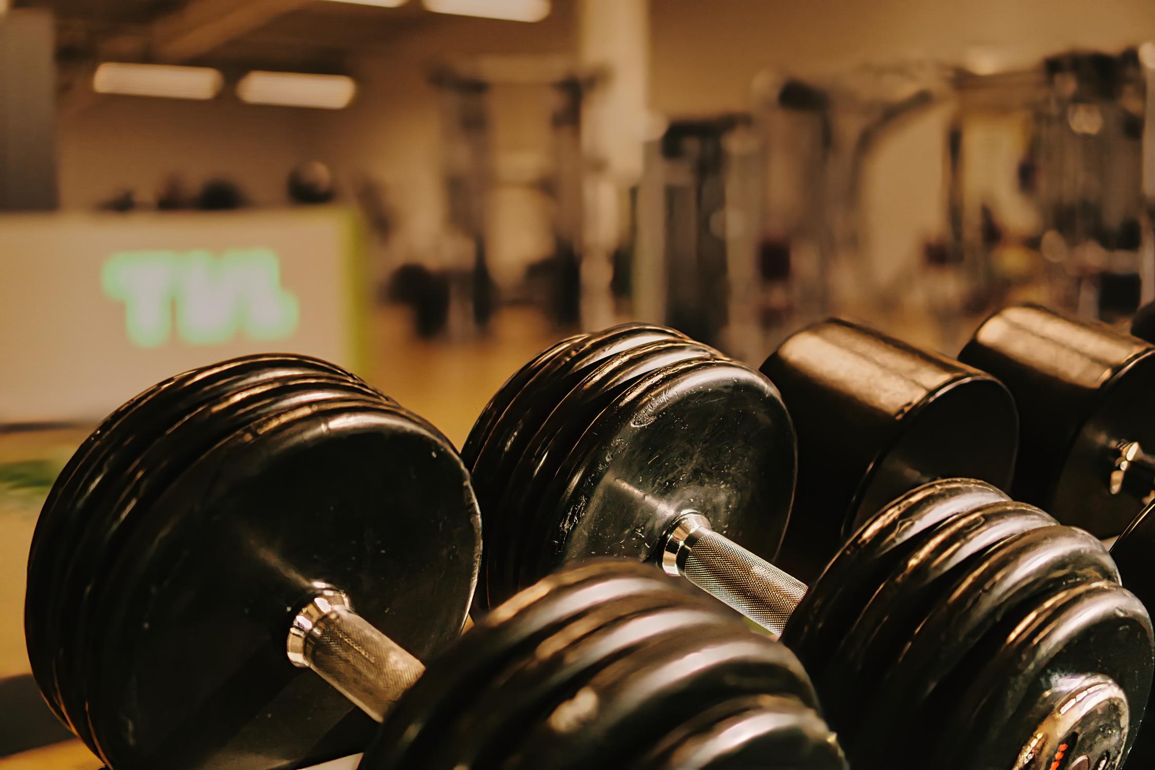 Nahaufnahme mehrerer schwarzer Kurzhanteln auf einem Fitnessstudio-Hintergrund mit Geräten und einer Beleuchtung.