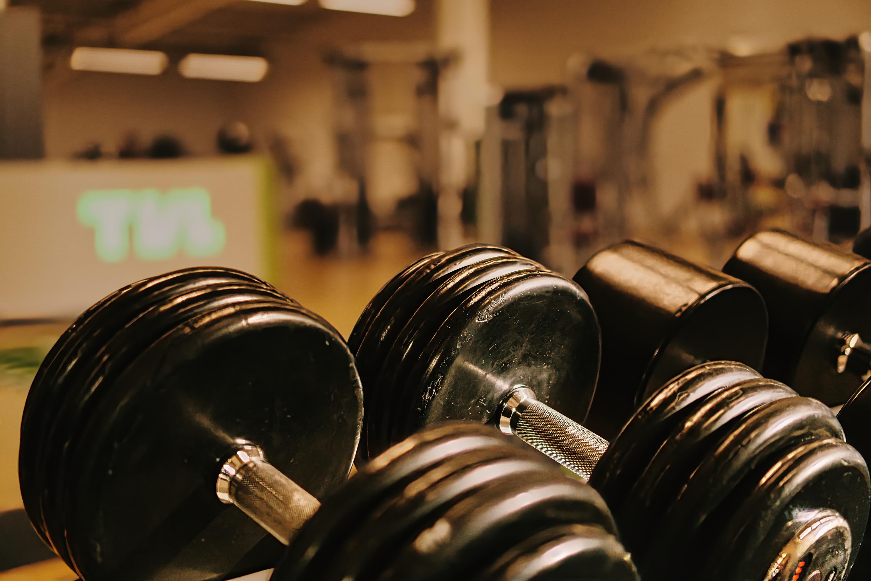 Nahaufnahme mehrerer schwarzer Kurzhanteln auf einem Fitnessstudio-Hintergrund mit Ger&auml;ten und einer Beleuchtung.