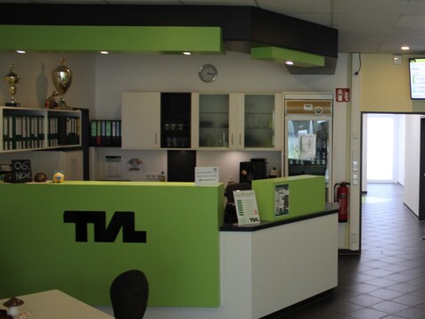 Lichtdurchfluteter Empfangsbereich mit gr&uuml;ner Theke, B&uuml;roschrank und digitalem Display an der Wand.