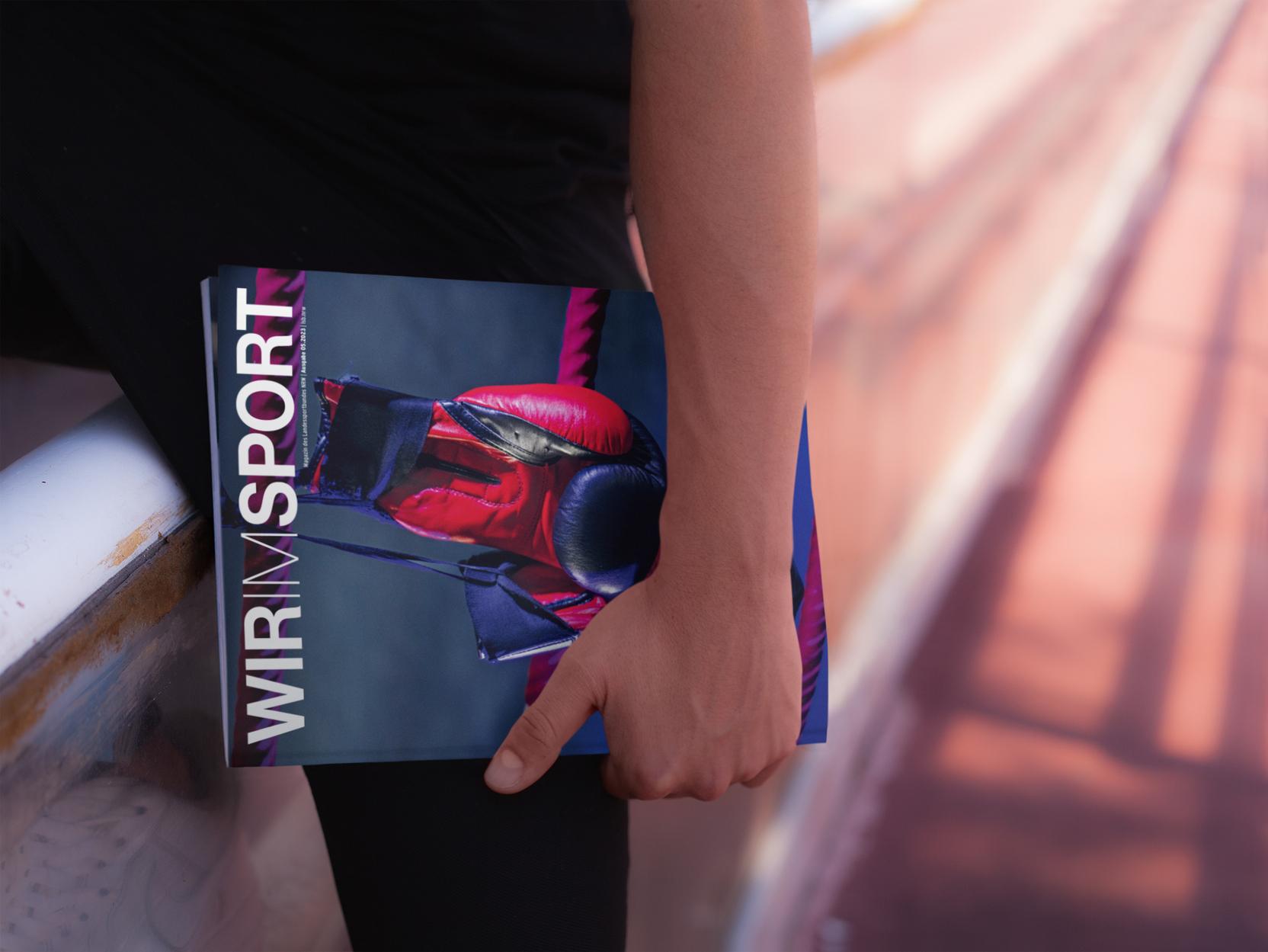 Hand h&auml;lt ein Magazin mit dem Titel &bdquo;WIR IM SPORT&ldquo; &uuml;ber einer Sportfl&auml;che. Die abgebildete Person tr&auml;gt sportliche Kleidung.