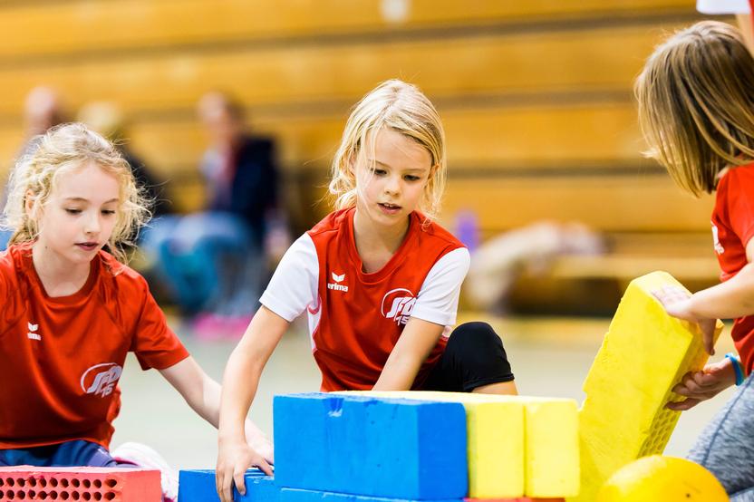 Drei Mädchen im roten Shirt bauen mit bunten Schaumstoffbausteinen in einer Turnhalle.