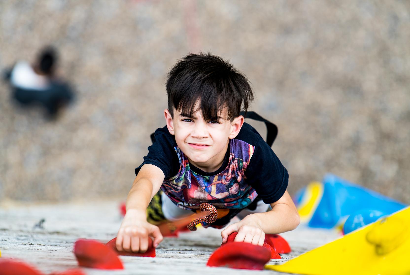 Junge klettert an einer Bouldering-Wand, greift nach einem roten Griff, mit angespannter Miene und sportlicher Kleidung.