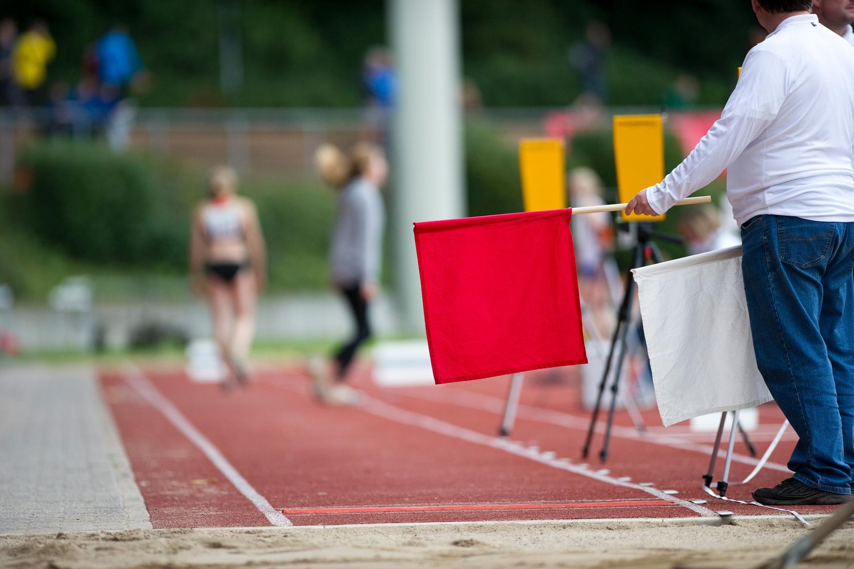 Ein Schiedsrichter hält eine rote Flagge neben einer Laufbahn, während Athleten im Hintergrund vorbeigehen.