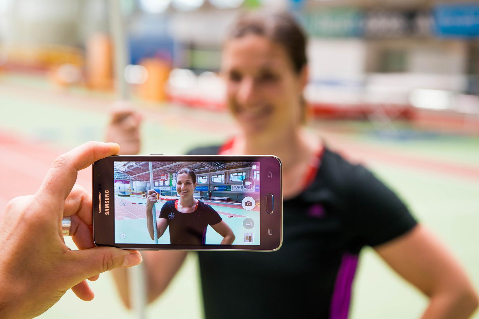 Hand hält ein Smartphone, das ein Porträt einer lächelnden Athletin mit Stab in einer Sporthalle zeigt.