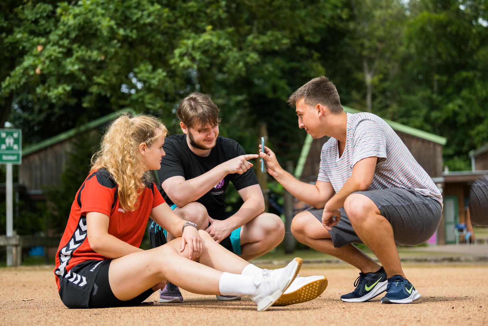 Drei Jugendliche sitzen auf einem Sandplatz und betrachten gemeinsam ein Smartphone. Bäume und Hütten im Hintergrund.