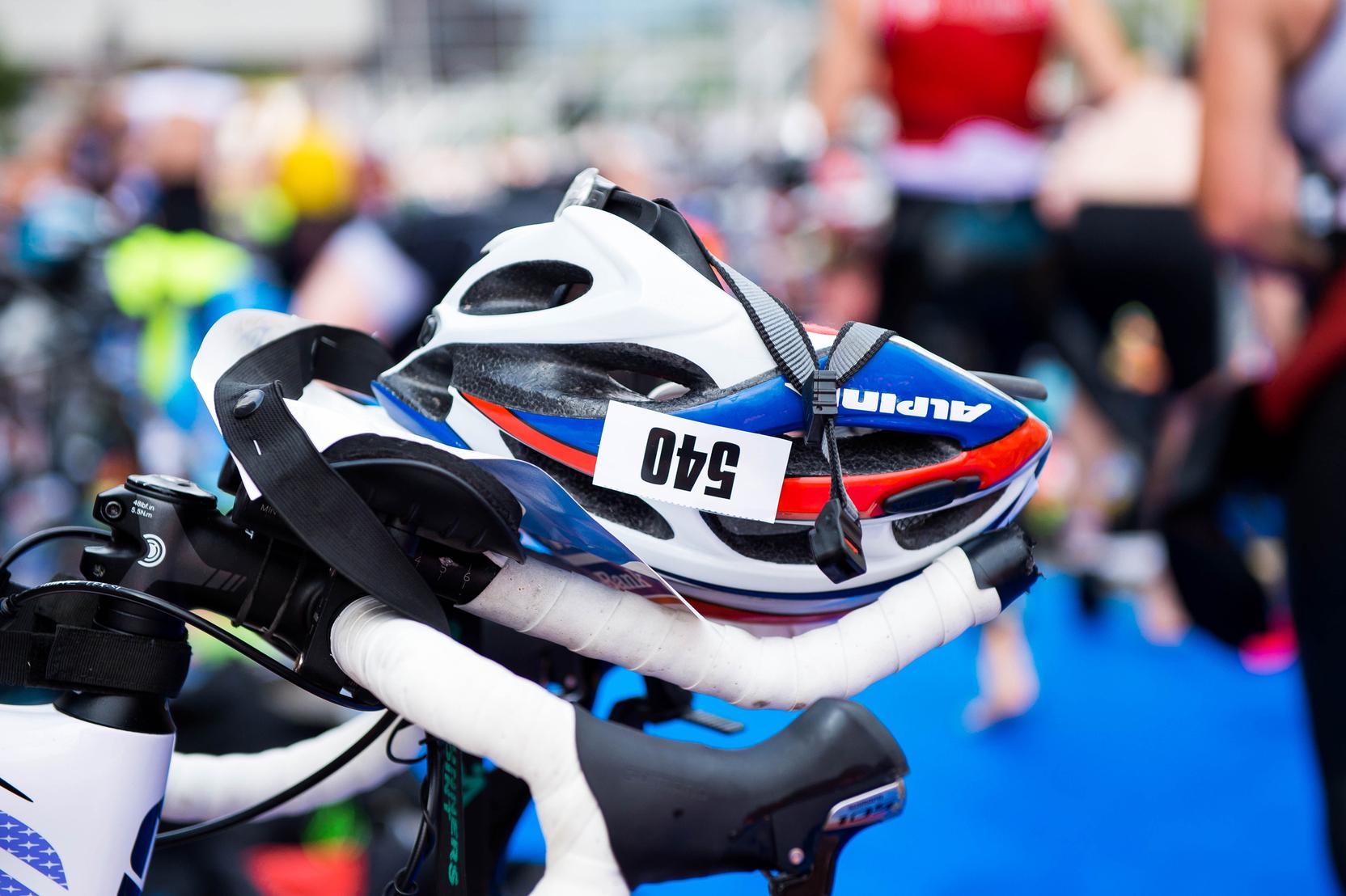 Nahaufnahme eines Fahrrads mit einem weißen Helm und einer Startnummer auf dem Lenker, Hintergrund unscharf.