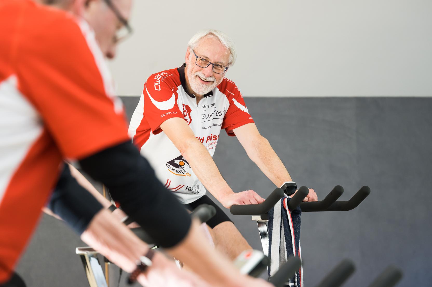 Älterer Mann mit Brille und grauen Haaren lächelt auf einem Spinning-Bike in einer Gruppe von Radfahrern.