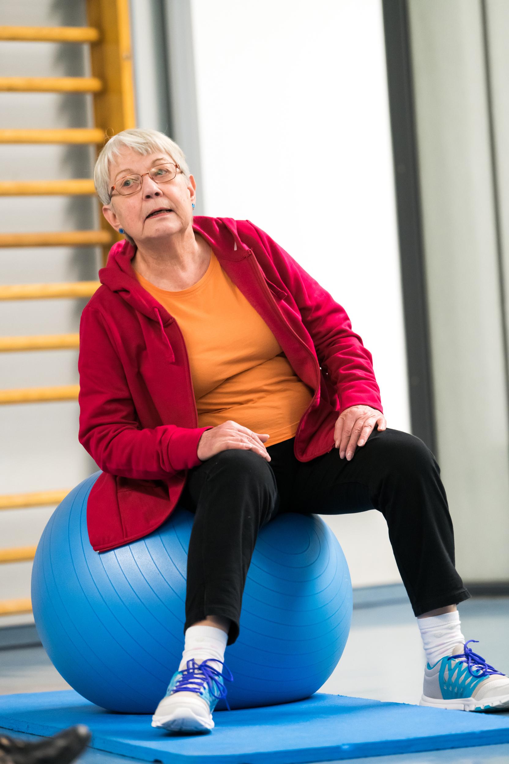 Ältere Frau in roter Jacke sitzt auf einem blauen Gymnastikball in einer Sporthalle.