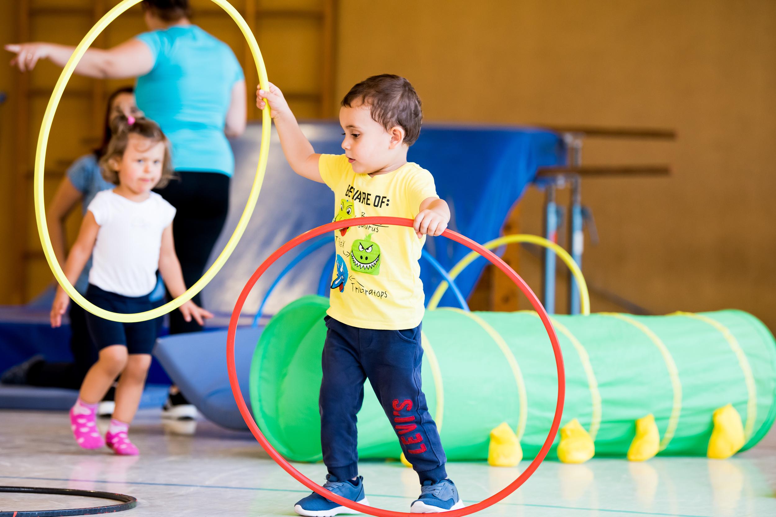 Zwei Kinder spielen in einer Turnhalle mit hula-hoops und einem grünen Tunnel. Ein Kind hält zwei Reifen in der Hand.